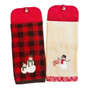 May include: Two hanging kitchen towels with red quilted tops and snowman buttons. One towel features a red and black buffalo plaid pattern with two embroidered snowmen. The other is cream-colored with a red snowflake border and a snowman with the text Let it Snow.