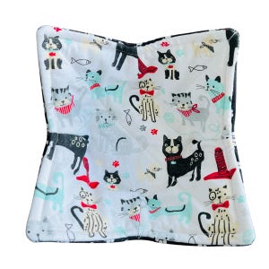 May include: A white fabric pot holder with a black border features a repeating pattern of black and white cats with red and blue accents. The cats are wearing bow ties and some have fish bones in their mouths.