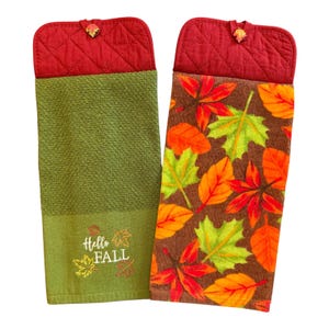 May include: Two hanging kitchen towels with quilted red tops and button closures. One towel is solid olive green with embroidered autumn leaves and the text Hello FALL. The other towel features a brown background with a pattern of orange, red, and green leaves.