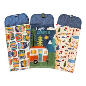 May include: Three hanging kitchen towels with quilted blue tops and button closures. The left towel features colorful sardine tin graphics. The center towel shows a camper, trees, and the text Happy Camper. The right towel displays Michigan motifs.