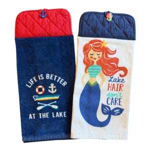 May include: Two hanging kitchen towels. One is navy blue with the text "LIFE IS BETTER AT THE LAKE" and nautical graphics. The other is white with a mermaid illustration and the text "Lake HAIR don't CARE". Both have a quilted top.