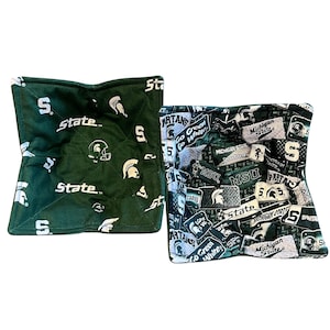 May include: Two green and white bowl cozies. One features the word "State" and the letter "S" with a Spartan helmet. The other has a collage of "Michigan State" logos, "Spartans", and "Go Green Go White" text.