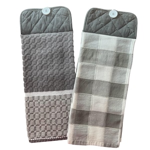 May include: Two gray kitchen towels with a quilted backing and a button closure. One towel has a waffle weave pattern and the other has a checkered pattern.