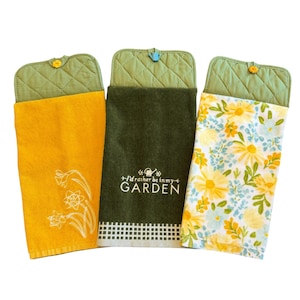 May include: Three hanging kitchen towels with green quilted tops and decorative buttons. One towel is yellow with a daffodil design, another is dark green with the text "I'd rather be in my GARDEN", and the third is white with yellow floral print.