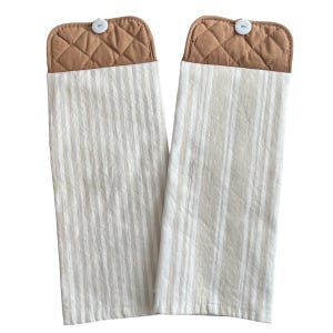 May include: Two hanging kitchen towels. The towels have a cream and white striped pattern with a tan quilted top and a white button. The towels are made of a soft, absorbent material.