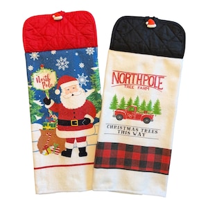 May include: Two hanging kitchen towels with quilted tops. The left towel features a red quilted top, a blue background with snowflakes, Santa Claus, and a North Pole sign. The right towel has a black quilted top, a white background, a red truck, and the text Northpole Tree Farm Christmas Trees This Way with a red and black buffalo plaid border.