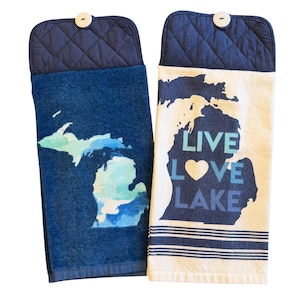 May include: Two hanging kitchen towels. One is dark blue with a watercolor map of Michigan. The other is cream-colored with a Michigan map and the words "LIVE LOVE LAKE" in blue. Both have a navy blue quilted top and a wooden button.