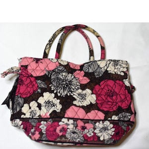 May include: A quilted fabric handbag featuring a dark brown background with a vibrant floral pattern in shades of hot pink, light pink, white, and black. The bag includes two matching quilted handles and a horizontal floral border at the base.