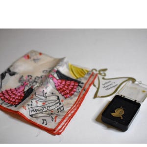 Puede incluir: Un pañuelo vintage con borde rojo y un diseño con bailarines con vestidos rosas. El texto "Arthur Murray" es visible. También hay un colgante dorado en una caja negra.