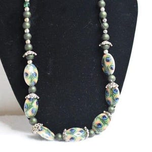 Puede incluir: Un collar de cuentas con cuentas ovaladas con un diseño de pluma de pavo real en azul, verde y melocotón. El collar tiene cuentas más pequeñas de color verde oliva y detalles plateados. El collar se muestra sobre un fondo negro.