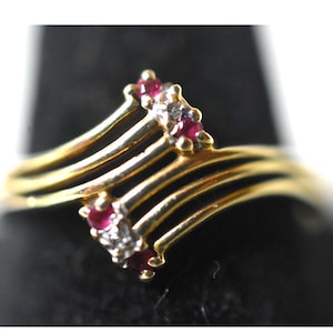 May include: A gold ring with a unique design. The ring features a series of parallel bands that curve upwards, adorned with small red gemstones and clear accent stones. The gemstones are set in a cluster at the top of the ring.