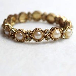 May include: Gold-toned stretch bracelet featuring faux pearls and small clear crystals. The bracelet has a textured, ornate design around each pearl and crystal setting. The pearls are cream-colored, and the crystals sparkle against the gold.