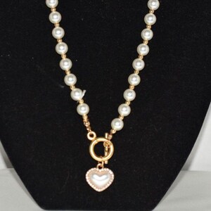 May include: A gold-toned necklace featuring a strand of white faux pearls. The necklace has a heart-shaped pendant with a pearl center and a gold-toned border. The necklace is displayed against a black background.