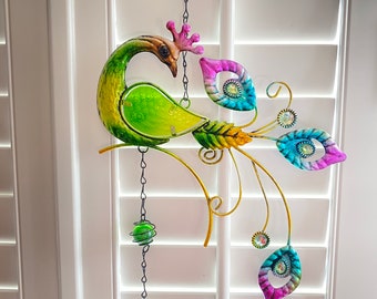 May include: A green and iridescent peacock wind chime with a bell hanging from the bottom. The peacock is perched on a branch with its tail feathers spread out. The feathers are decorated with colourful jewels.