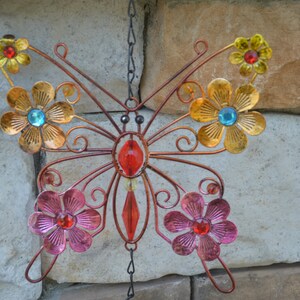 May include: A decorative metal butterfly with copper-coloured wings and body, adorned with colourful flowers and gemstones. The flowers are in shades of gold, pink, and yellow, with blue and red gemstone centres. The butterfly hangs from a chain.