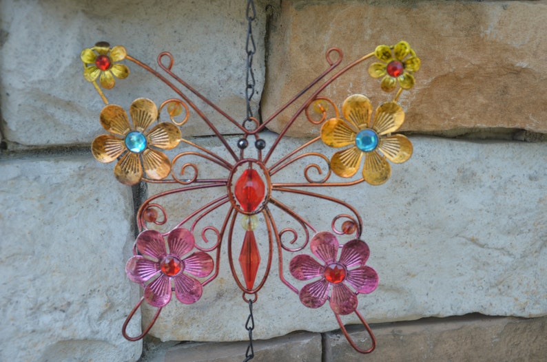 May include: A decorative metal butterfly with copper-coloured wings and body, adorned with colourful flowers and gemstones. The flowers are in shades of gold, pink, and yellow, with blue and red gemstone centres. The butterfly hangs from a chain.