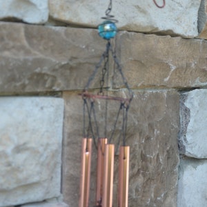 May include: A decorative wind chime with copper-coloured tubes and a butterfly-shaped top. The butterfly has pink flower accents and a red centre. A blue glass bead hangs above the chimes, suspended by a chain.