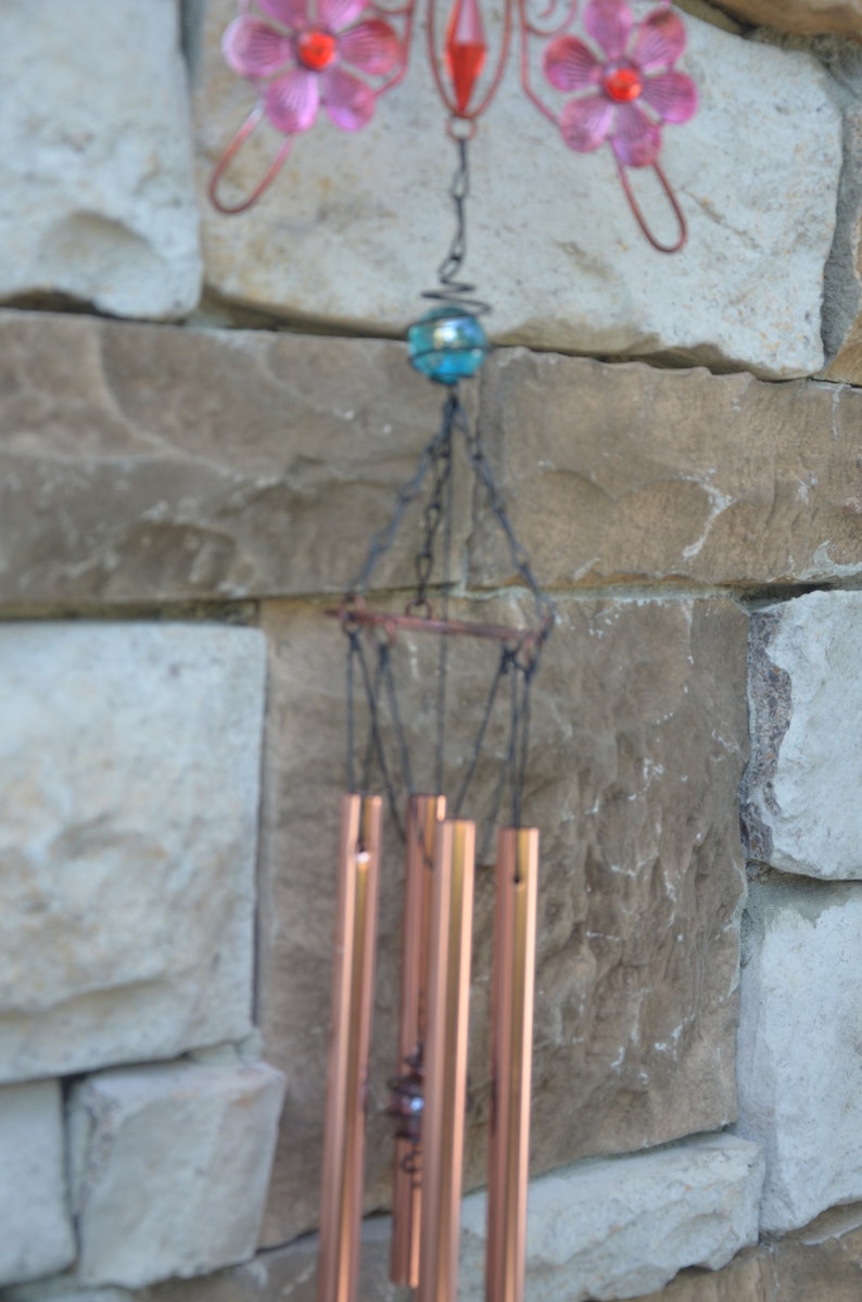 May include: A decorative wind chime with copper-coloured tubes and a butterfly-shaped top. The butterfly has pink flower accents and a red centre. A blue glass bead hangs above the chimes, suspended by a chain.