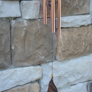 May include: Copper-toned wind chimes with a leaf-shaped charm suspended from a black string. The chimes are set against a backdrop of a stone wall with varying shades of beige and grey.