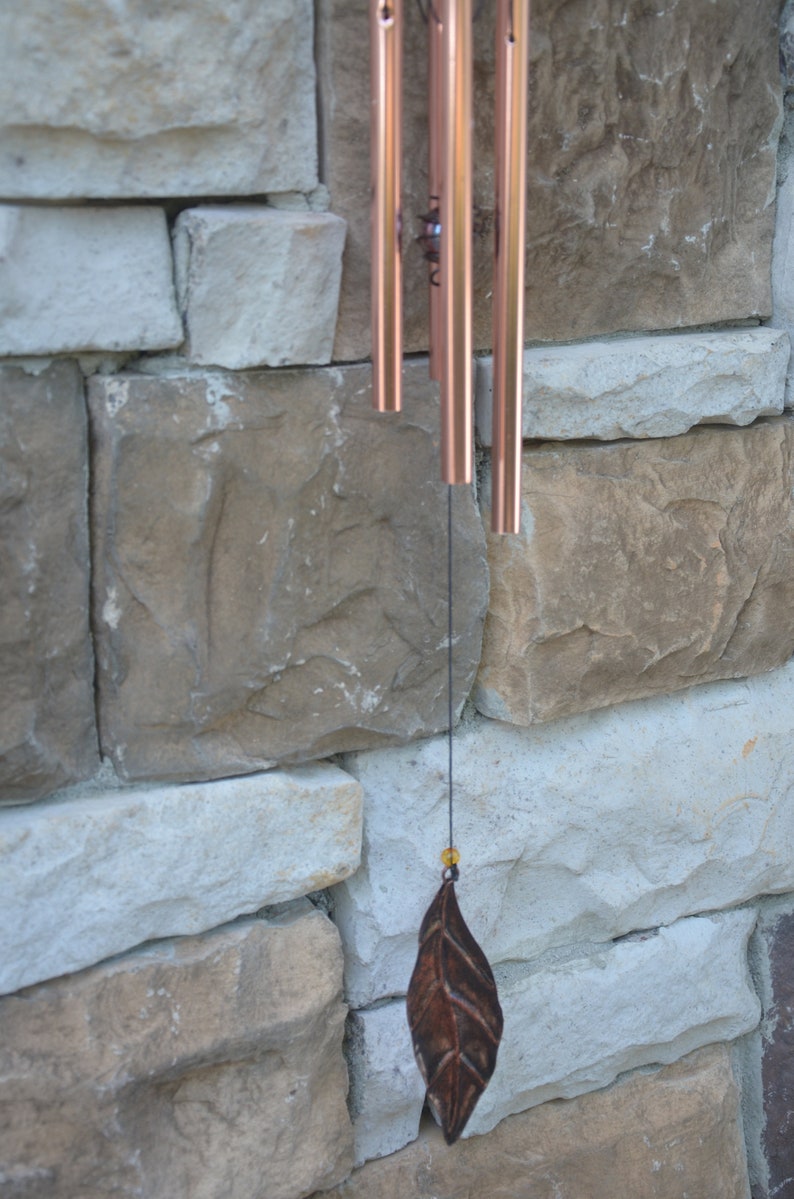 May include: Copper-toned wind chimes with a leaf-shaped charm suspended from a black string. The chimes are set against a backdrop of a stone wall with varying shades of beige and grey.