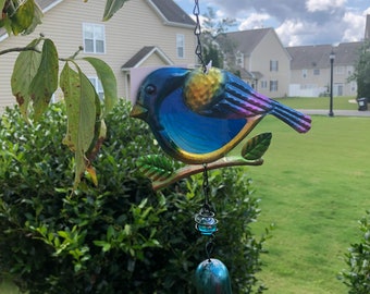 May include: A blue, yellow and purple bird wind chime with a bell hanging from a chain. The bird is perched on a branch with green leaves. The wind chime is hanging from a tree branch.