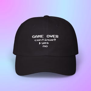 May include: Black baseball cap with white text that reads "GAME OVER continue?" with "yes" and "no" options. The cap has a curved brim and a structured crown, suitable for casual wear. The background is a gradient of pink and blue.