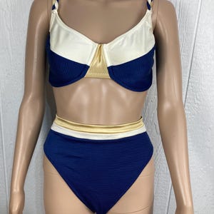 May include: Two-piece swimsuit in navy blue, cream, and gold. The top has a cream-colored upper section and navy blue lower section with gold accents. The high-waisted bottoms are navy blue with gold and cream trim. Braided straps.