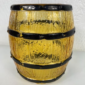 May include: A decorative barrel-shaped container with a textured, golden-yellow glass body. The barrel is accented with black bands and rivets, giving it a rustic appearance. The container is designed to resemble a miniature wooden barrel.
