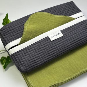Peut inclure: Un ensemble de deux torchons de cuisine en tissu gaufré, un gris et un vert. Le torchon gris porte une étiquette blanche avec le texte "La Girafe". Les torchons sont pliés et empilés l'un sur l'autre.
