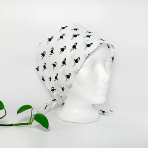 Op de afbeelding: Witte chirurgische scrub-muts met een zwarte flamingo print. De muts heeft banden die aan de achterkant van de nek kunnen worden vastgebonden.