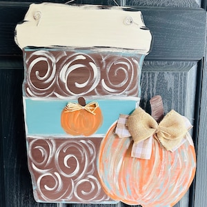 May include: A wooden coffee cup door hanger with a brown and white swirl design. The cup has a blue band with a small orange pumpkin. A larger orange pumpkin with a burlap bow is attached to the side of the cup.