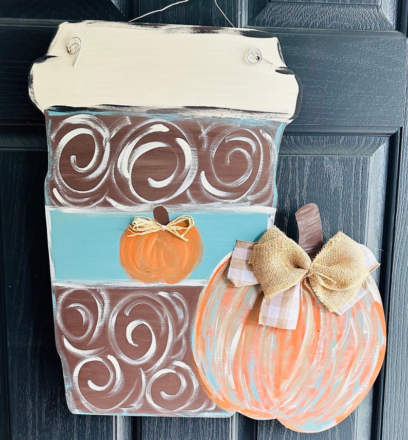 May include: A wooden coffee cup door hanger with a brown and white swirl design. The cup has a blue band with a small orange pumpkin. A larger orange pumpkin with a burlap bow is attached to the side of the cup.