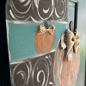 May include: A wooden door hanger shaped like a coffee cup with a brown and white swirl pattern. The cup has a teal blue band and two pumpkins with bows on the side. One pumpkin has a brown bow and the other has a gold bow.