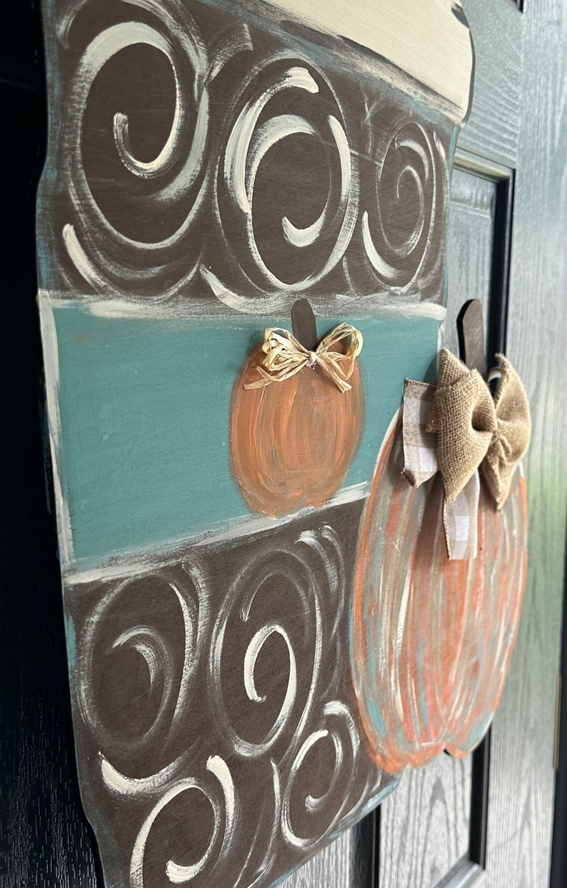 May include: A wooden door hanger shaped like a coffee cup with a brown and white swirl pattern. The cup has a teal blue band and two pumpkins with bows on the side. One pumpkin has a brown bow and the other has a gold bow.