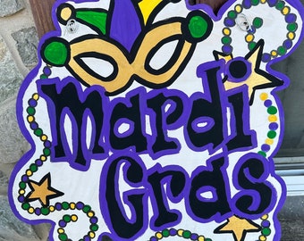 May include: A purple, yellow, and green Mardi Gras sign with a jester's hat, mask, and beads. The sign says "Mardi Gras" in black letters.