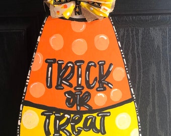 May include: A wooden candy corn door hanger with a yellow and orange polka dot pattern. The words "Trick or Treat" are written in black on the candy corn. A yellow and white bow is attached to the top of the candy corn.