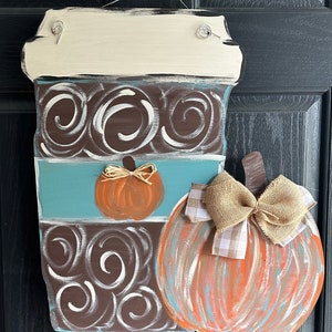 May include: A wooden coffee cup door hanger with a brown and white swirl design, a blue stripe, and a small orange pumpkin. A larger orange pumpkin with a brown stem and a burlap bow is attached to the side of the coffee cup.