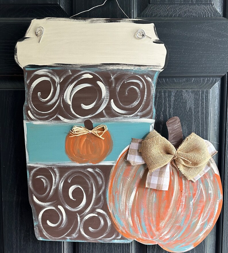 May include: A wooden coffee cup door hanger with a brown and white swirl design, a blue stripe, and a small orange pumpkin. A larger orange pumpkin with a brown stem and a burlap bow is attached to the side of the coffee cup.