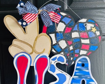 May include: A wooden door hanger with a peace sign, a heart, and the letters "USA" painted in red, white, and blue. The heart is decorated with a checkered pattern and a star. The peace sign is painted in a light brown color. The letters are painted in a bold, stylized font. The hanger is decorated with a red, white, and blue bow.