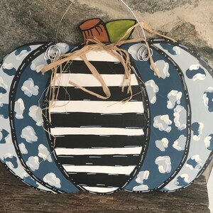 Puede incluir: Un colgador de puerta de calabaza de madera pintado con un patr&oacute;n de rayas blancas y negras con detalles de estampado de leopardo azul y blanco. La calabaza tiene un tallo de paja y est&aacute; lista para colgar.