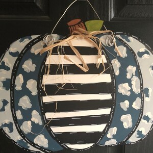 Puede incluir: Un colgador de puerta de calabaza pintado en azul y blanco con un centro a rayas en blanco y negro. La calabaza tiene un dise&ntilde;o de estampado de leopardo blanco y azul en los lados.