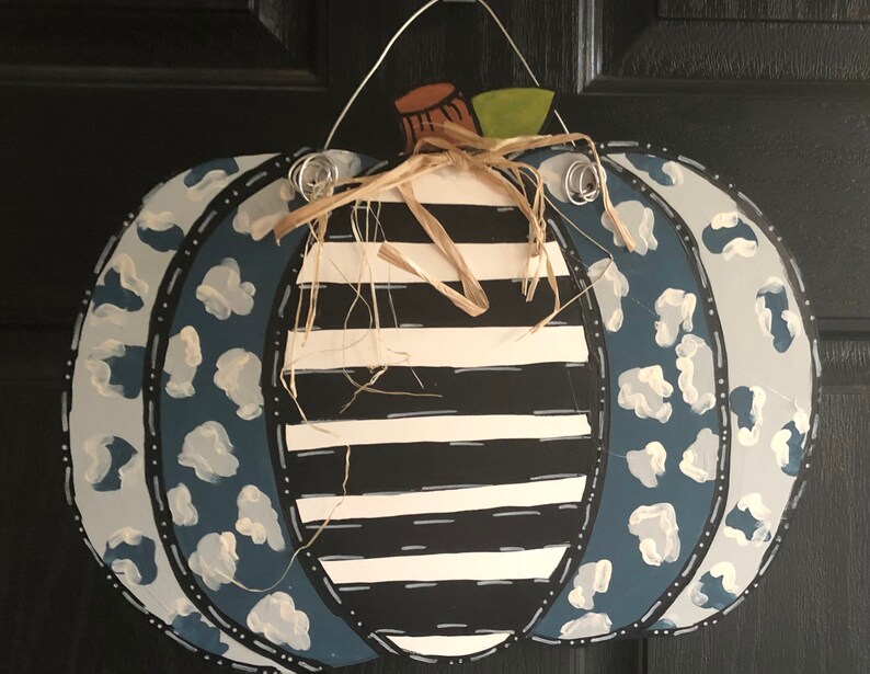 Puede incluir: Un colgador de puerta de calabaza pintado en azul y blanco con un centro a rayas en blanco y negro. La calabaza tiene un dise&ntilde;o de estampado de leopardo blanco y azul en los lados.
