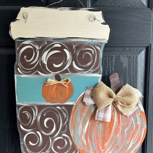 May include: A wooden coffee cup door hanger with a brown and white swirl design, a blue stripe, and a small orange pumpkin. A large orange pumpkin with a brown burlap bow is attached to the side of the coffee cup.