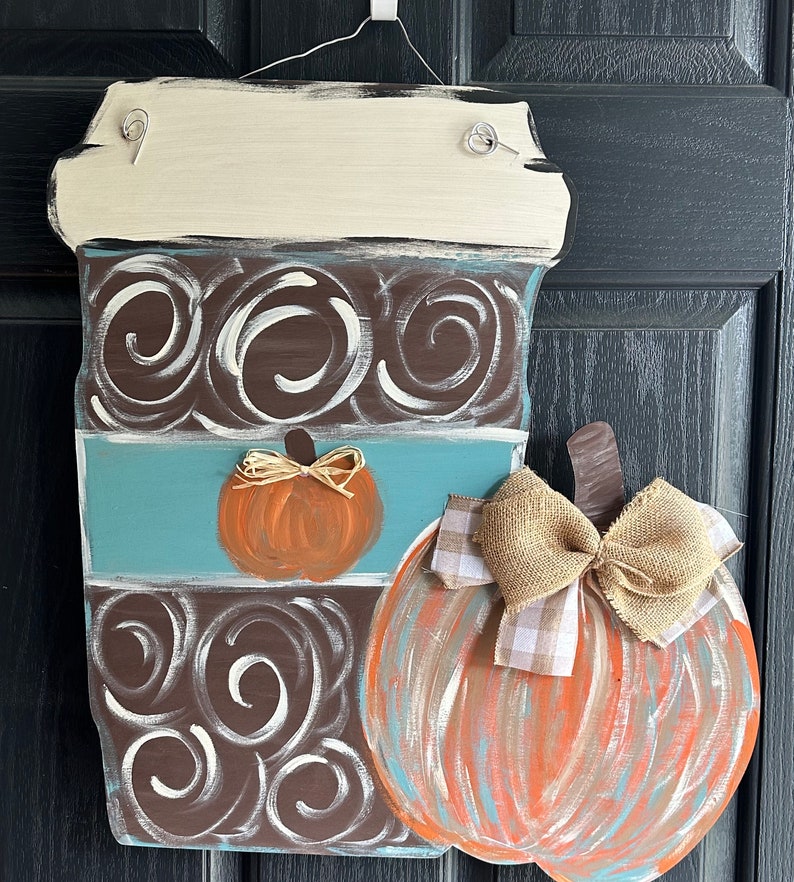 May include: A wooden coffee cup door hanger with a brown and white swirl design, a blue stripe, and a small orange pumpkin. A large orange pumpkin with a brown burlap bow is attached to the side of the coffee cup.