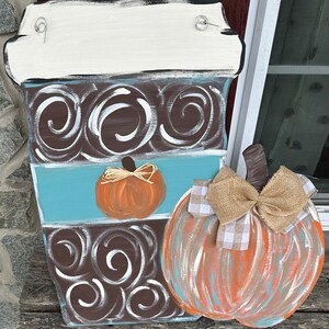 May include: A wooden coffee cup door hanger with a brown and white swirl pattern, a blue stripe, and a small orange pumpkin. A large orange pumpkin with a burlap bow and a white and brown checkered bow is attached to the side of the coffee cup.