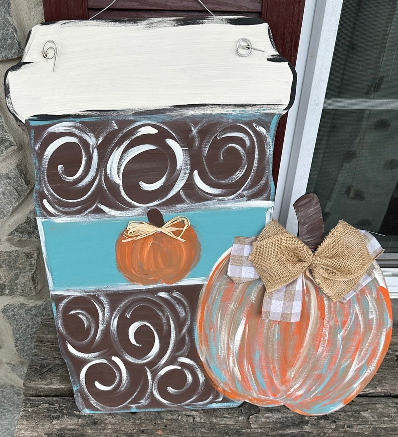 May include: A wooden coffee cup door hanger with a brown and white swirl pattern, a blue stripe, and a small orange pumpkin. A large orange pumpkin with a burlap bow and a white and brown checkered bow is attached to the side of the coffee cup.