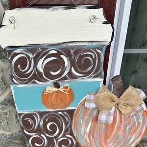 May include: A wooden coffee cup door hanger with a brown and white swirl design, a blue stripe, and a small orange pumpkin. A larger pumpkin with a brown stem and a burlap bow is attached to the side of the coffee cup.