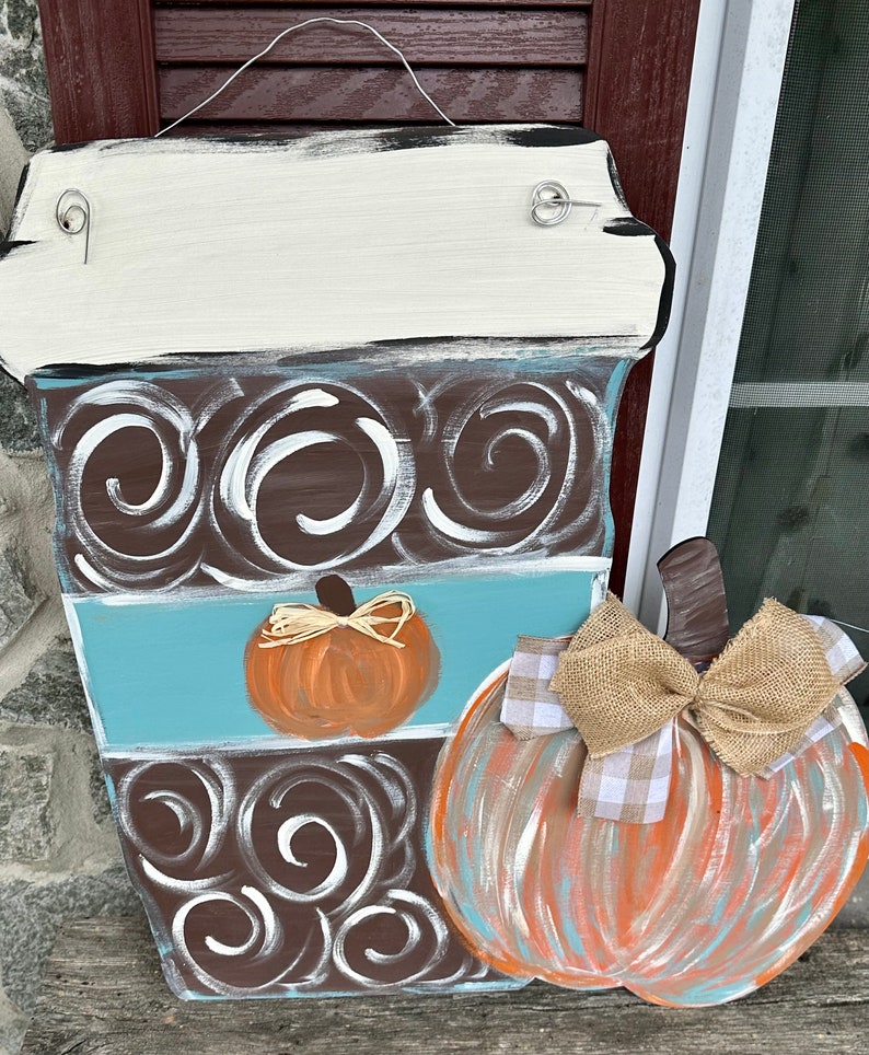 May include: A wooden coffee cup door hanger with a brown and white swirl design, a blue stripe, and a small orange pumpkin. A larger pumpkin with a brown stem and a burlap bow is attached to the side of the coffee cup.