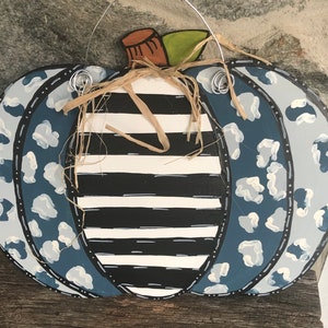 Puede incluir: Un colgador de puerta de calabaza de madera pintado con un patr&oacute;n de rayas blancas y negras con detalles de estampado de leopardo azul y blanco. La calabaza tiene un tallo de paja y est&aacute; lista para colgar.