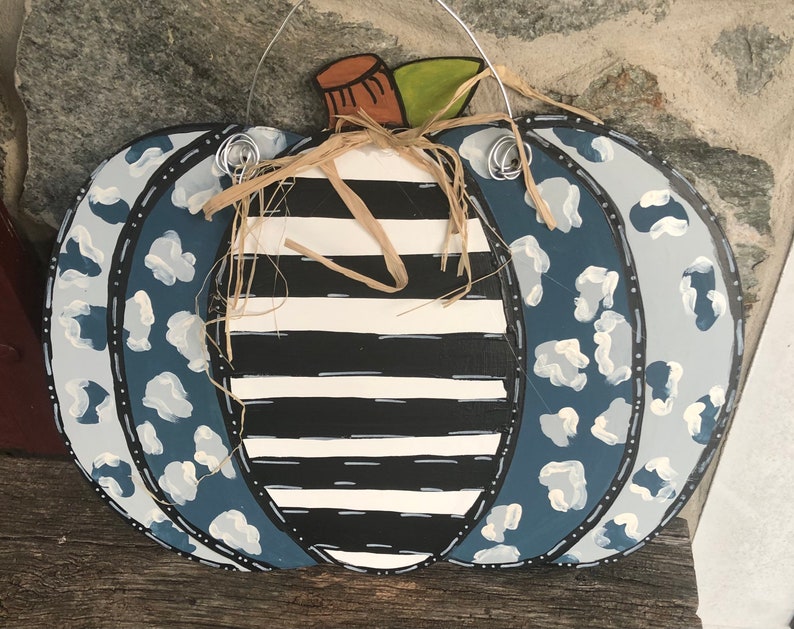 Puede incluir: Un colgador de puerta de calabaza de madera pintado con un patr&oacute;n de rayas blancas y negras con detalles de estampado de leopardo azul y blanco. La calabaza tiene un tallo de paja y est&aacute; lista para colgar.